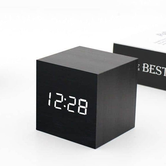Black cube-shaped digital clock on a white surface with a plant and book in the background.