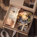 Gift set with a rose gold bottle and a small bouquet of flowers in an open box on a wooden surface.