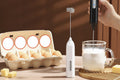 Handheld milk frother with a glass of milk being frothed on a wooden surface.