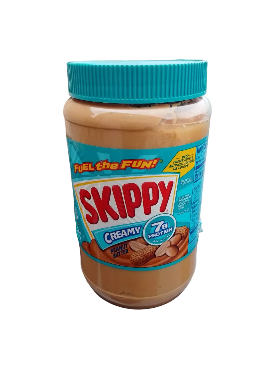 A jar of Skippy Creamy Peanut Butter Protein spread with a bright blue lid, sitting on a dark reflective surface with a blurred outdoor background.